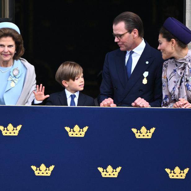 Königin Silvia, Prinz Oscar, Prinz Daniel und Kronprinzessin Victoria stehen auf einem Balkon mit königlichem Wappen und winken der Menge zu.