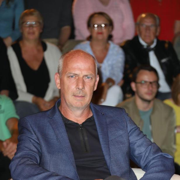 Mario Basler sitzt mit verschränkten Beinen in einem Sessel, im Hintergrund sind mehrere Zuschauer zu sehen.