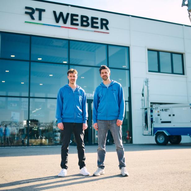 Zwei Männer in blauen Kapuzenjacken stehen vor einem modernen Firmengebäude mit der Aufschrift „RT Weber“.