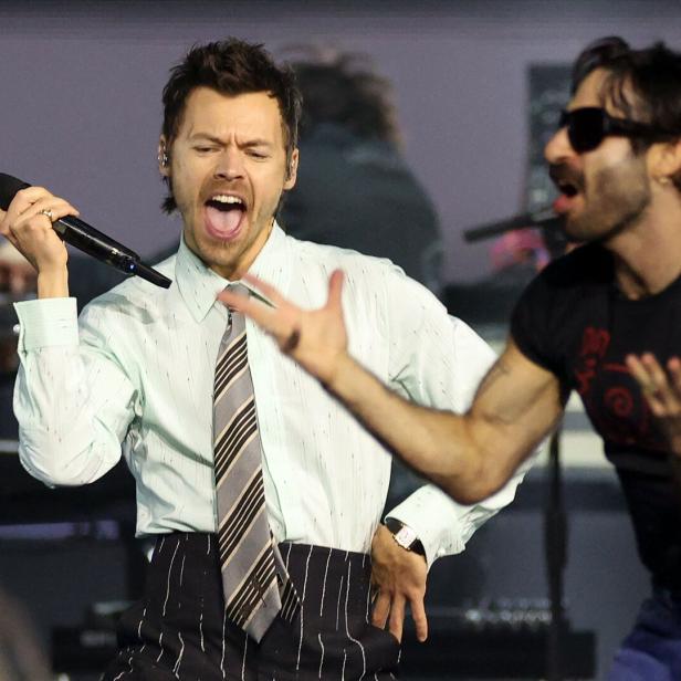 Harry Styles singt mit einem Mikrofon auf einer Bühne, während ein Mann mit Sonnenbrille neben ihm gestikuliert.