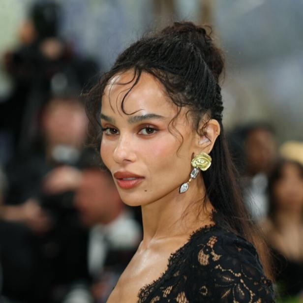 Zoë Kravitz trägt ein schwarzes Spitzenoberteil und auffällige goldene Ohrringe vor einem unscharfen Hintergrund.