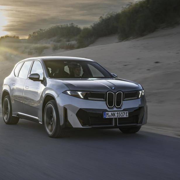 Ein silberner BMW Neue Klasse iX3 fährt auf einer Straße an einer Dünenlandschaft vorbei.