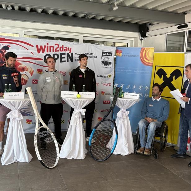Vier Männer stehen und einer sitzt im Rollstuhl bei einer Pressekonferenz mit Tennisbezug vor Sponsorenwänden und großen Tennisschlägern.