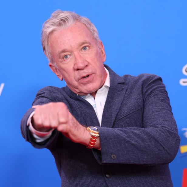 Tim Allen posiert vor einer blauen Wand mit Disney-Pixar-Logos und streckt seine Faust in die Kamera.