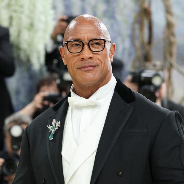 Dwayne Johnson trägt einen eleganten schwarzen Smoking mit weißer Weste, Fliege, Brille und einer auffälligen Brosche vor unscharfem Hintergrund.