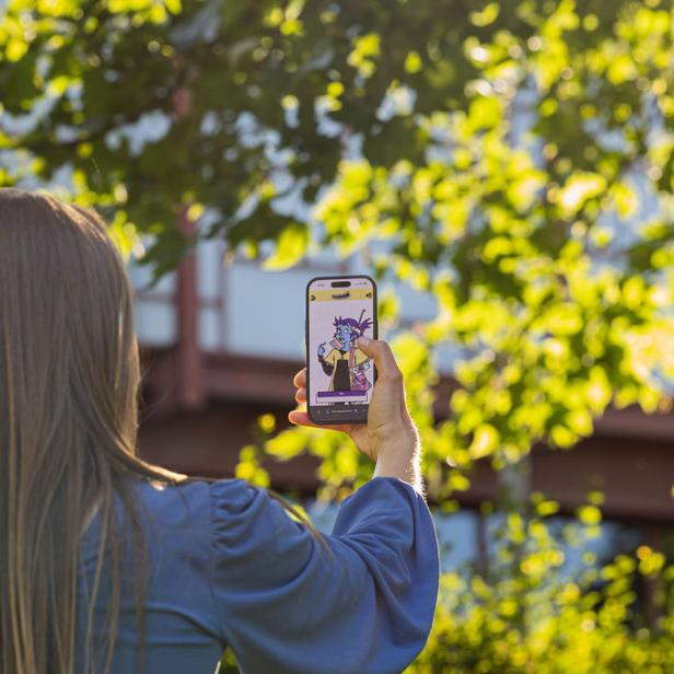 Eine Person mit langen Haaren hält ein Smartphone hoch, auf dessen Bildschirm eine bunte Comicfigur zu sehen ist, umgeben von grünen Bäumen.