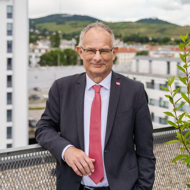 Markus Marterbauer im Anzug mit roter Krawatte steht auf einer Terrasse, im Hintergrund sind Gebäude und grüne Hügel zu sehen.