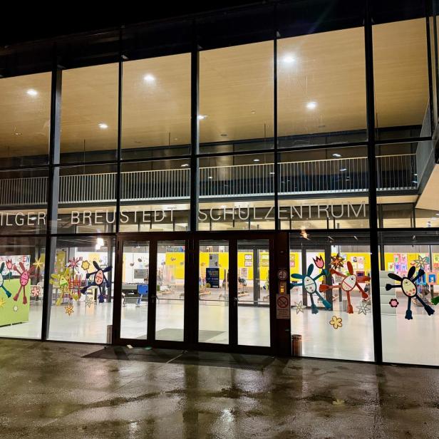 Großes, beleuchtetes Schulgebäude mit Glasfassade, bunten Kinderzeichnungen und dem Schriftzug „Bilger Breustedt Schulzentrum“.