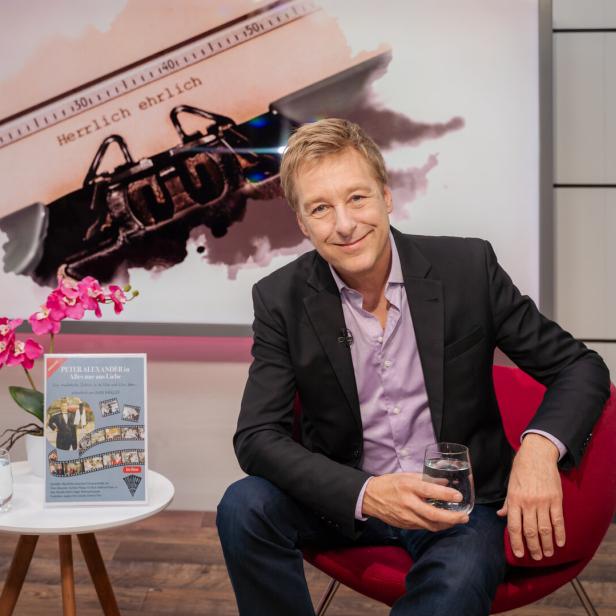 Max Müller sitzt in einem roten Sessel vor einem Plakat über Peter Alexander und hält ein Glas Wasser in der Hand.