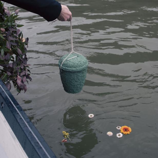 Eine Person hält eine grüne Urne über das Wasser, während Blumen auf der Wasseroberfläche treiben.