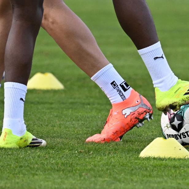 Zwei Fußballspieler mit bunten Schuhen stehen auf dem Rasen, einer hält einen Ball zwischen gelben Markierungshütchen.