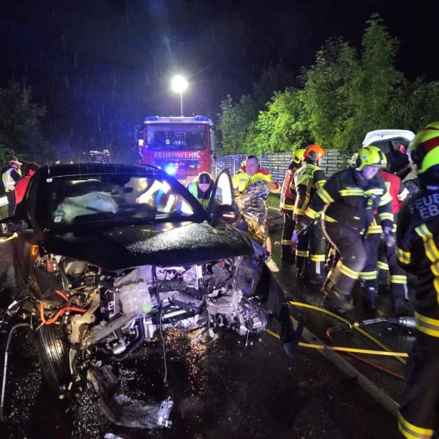 Ein schwer beschädigtes Auto bei Nacht, umgeben von Feuerwehrleuten und Rettungskräften im Einsatz.