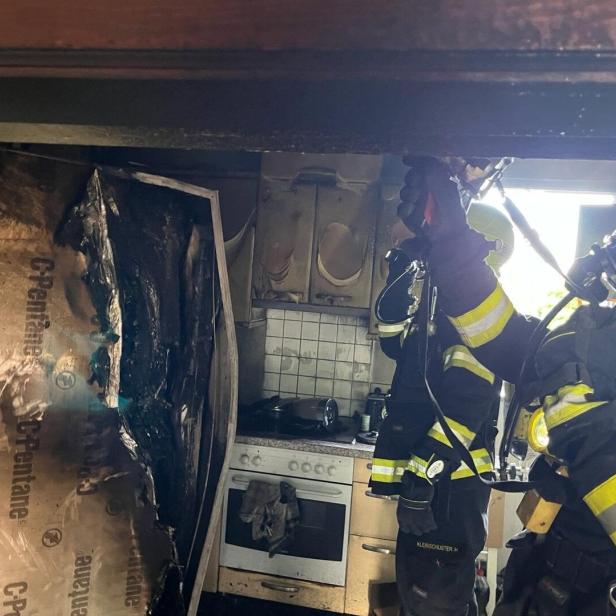 Zwei Feuerwehrleute in Schutzkleidung löschen einen Brand in einer stark beschädigten Küche mit verbrannten Geräten und Möbeln.