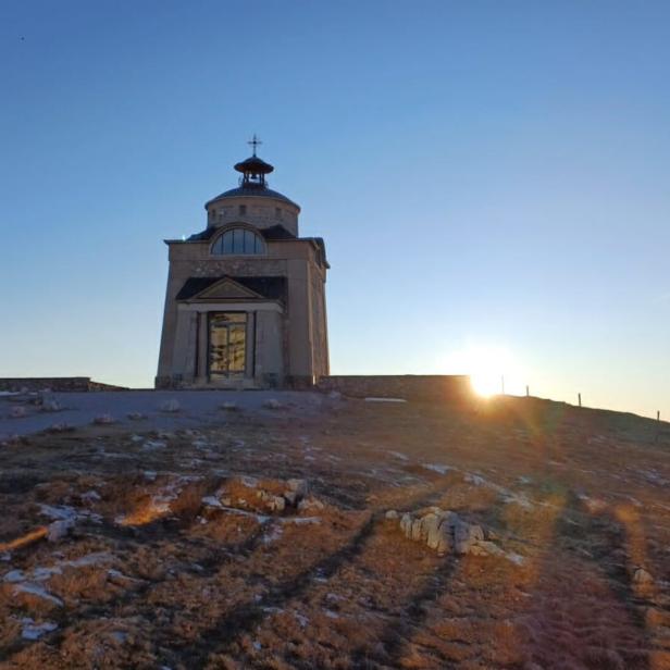 Kleine Kapelle auf einem Hügel bei Sonnenaufgang, mit klarem Himmel und spärlicher Vegetation im Vordergrund.