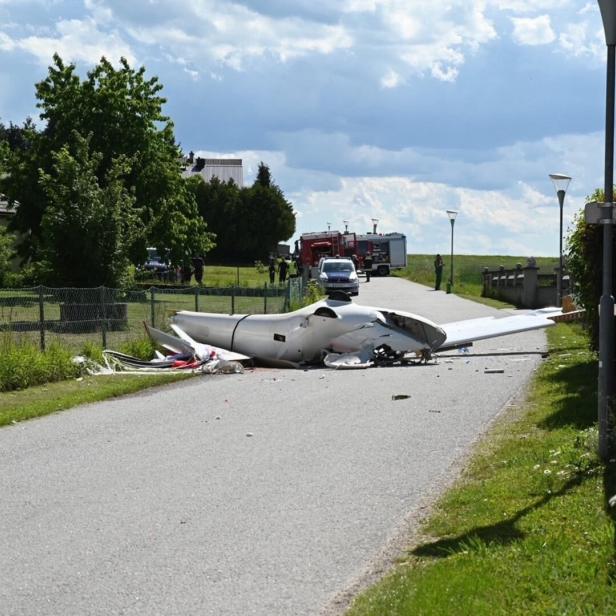 Ein abgestürztes Kleinflugzeug liegt zerstört auf einer Landstraße, Einsatzfahrzeuge und Einsatzkräfte im Hintergrund.
