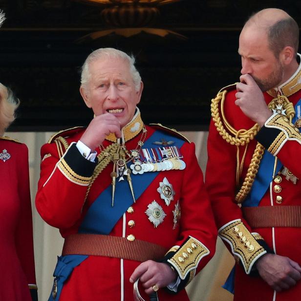 König Charles III., Königin Camilla und Prinz William in prunkvollen roten Uniformen auf einem Balkon.