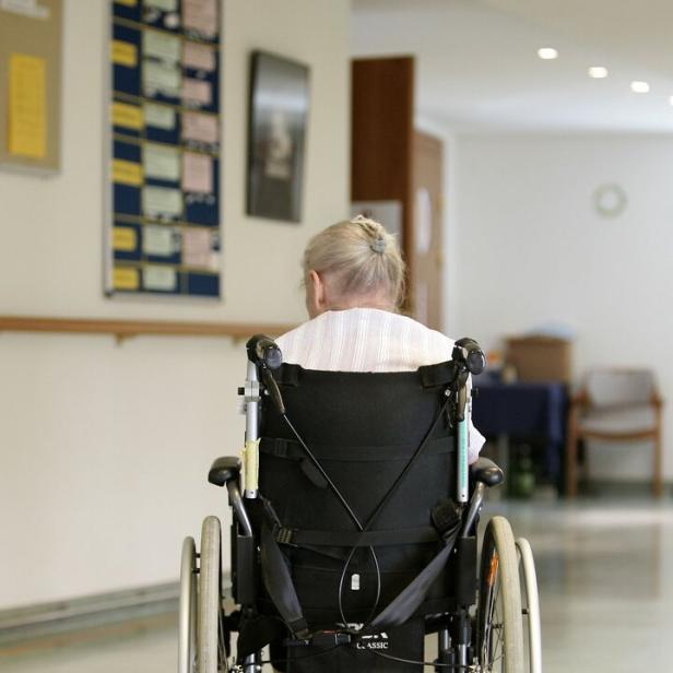 Eine ältere Person mit grauen Haaren sitzt im Rollstuhl auf einem Flur mit hellen Wänden und einigen Stühlen.