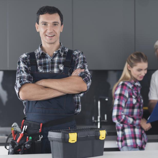Ein Handwerker steht mit verschränkten Armen und Werkzeugtasche im Vordergrund, während im Hintergrund zwei Personen ein Angebot besprechen.