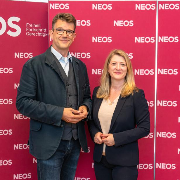 Ein Mann und eine Frau stehen lächelnd vor einer roten Wand mit dem wiederholten Logo der Partei NEOS.