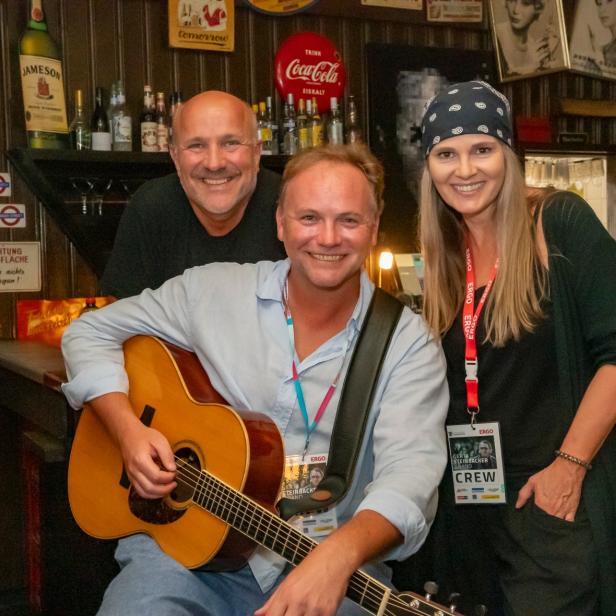 Drei fröhliche Menschen in einer Bar, einer hält eine Gitarre, die anderen stehen daneben und lächeln.