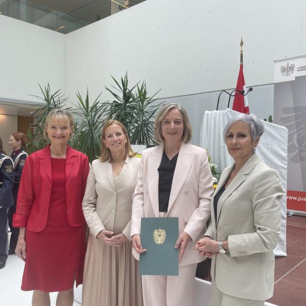 Vier Frauen in eleganter Kleidung stehen lächelnd nebeneinander, eine hält eine Urkunde mit österreichischem Wappen.