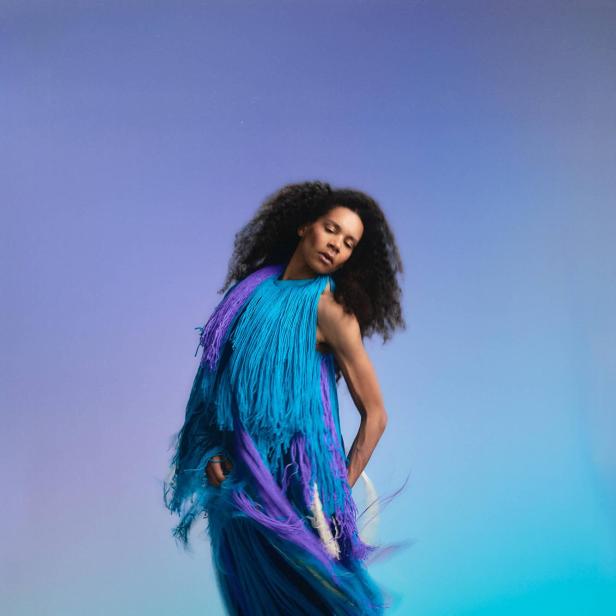 Rebecca Horner trägt ein fließendes Fadenkleid von Franz Josef Baur vor blauem Studiohintergrund.