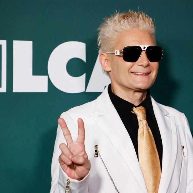 Corey Feldman mit blonden Haaren und Sonnenbrille trägt einen weißen Anzug vor einem Hintergrund mit dem Logo der The Bernheim Group.