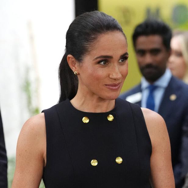 Meghan Markle trägt ein ärmelloses schwarzes Kleid mit goldenen Knöpfen und hat ihre Haare zu einem Pferdeschwanz zurückgebunden.
