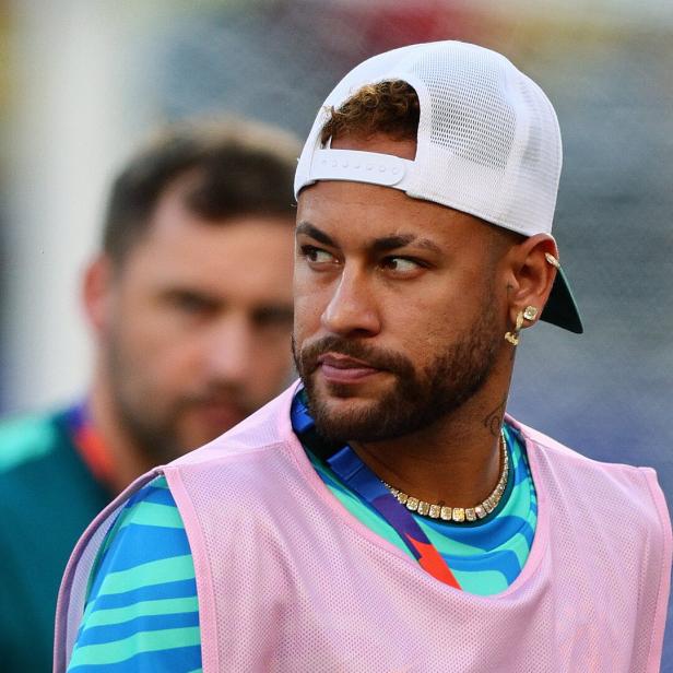 Neymar trägt eine weiße Kappe und ein pinkes Überziehshirt vor einem unscharfen Hintergrund.