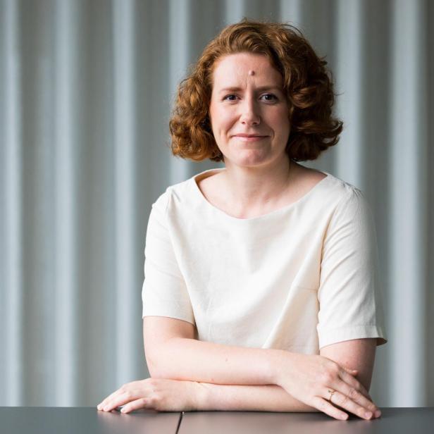 Eine Frau mit rötlichem, lockigem Haar und einem hellen Oberteil sitzt vor einem grauen, gestreiften Hintergrund.