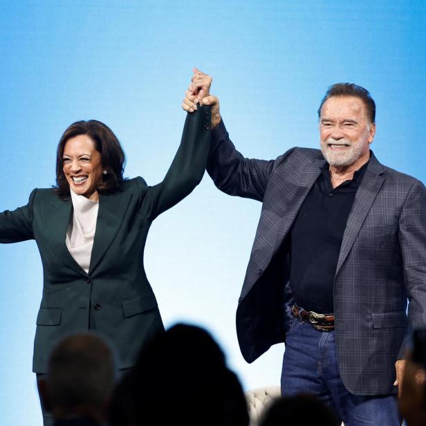 Kamala Harris und Arnold Schwarzenegger stehen lächelnd vor einem blauen Hintergrund und halten gemeinsam die Hände in die Höhe.
