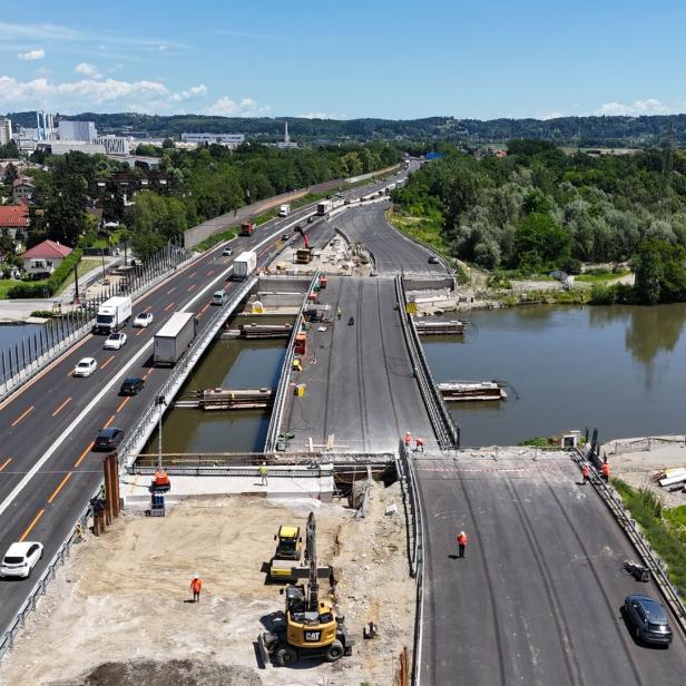 Luftaufnahme einer Brückenbaustelle über einen Fluss mit Autos, Baufahrzeugen und Stadt im Hintergrund.