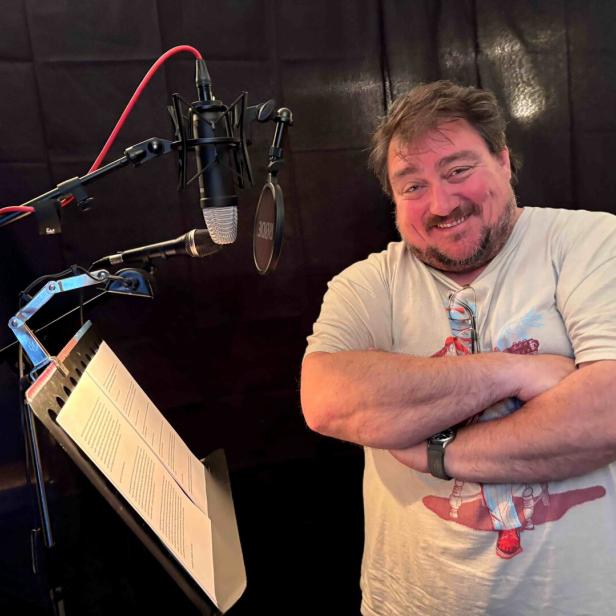 Ein lächelnder Mann steht vor Mikrofonen und einem Notenständer in einem Tonstudio.