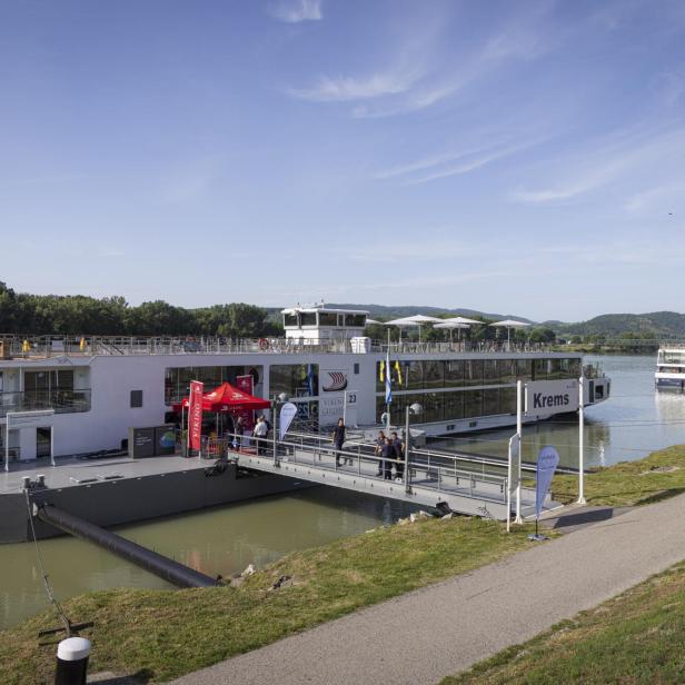 Anlegestelle am Fluss mit zwei Kreuzfahrtschiffen, Steg und Passagieren vor grünen Hügeln unter blauem Himmel.