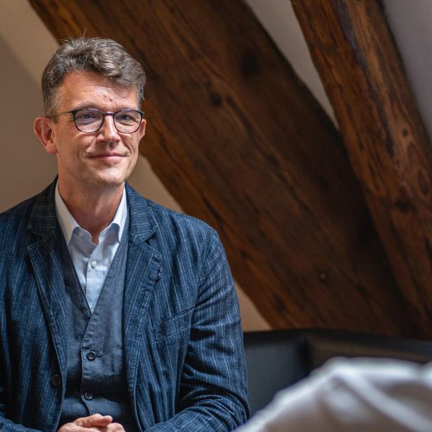 Ein Mann mit Brille und blauem Sakko sitzt in einem Gesprächsraum und blickt sein Gegenüber an.