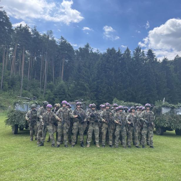 Soldaten mit Waffen posieren vor getarnten Militärfahrzeugen auf einer Wiese.