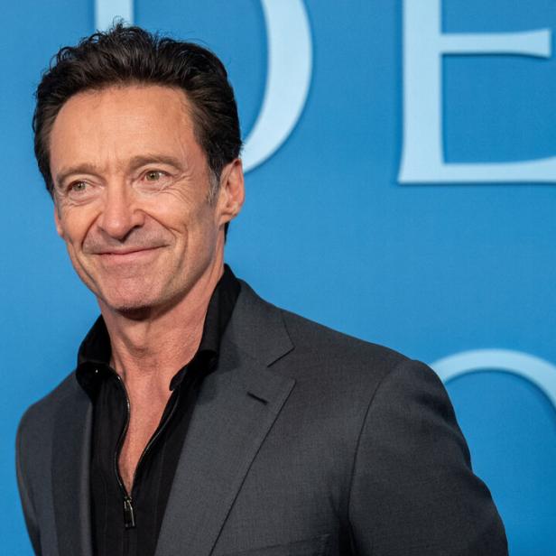 Hugh Jackman lächelt vor einem blauen Hintergrund in einem grauen Sakko und einem schwarzen Hemd.