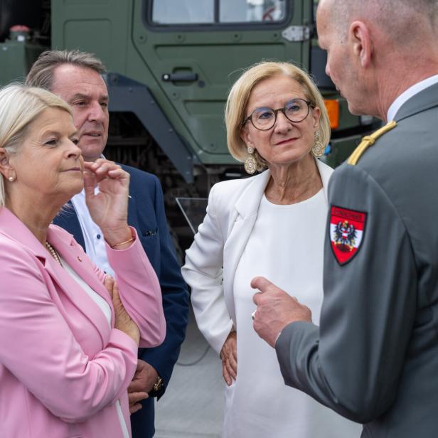 Vier Personen sprechen vor einem Militärfahrzeug, darunter eine Frau in Pink und ein Offizier in Uniform.
