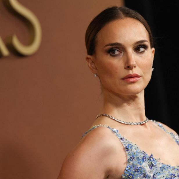 Natalie Portman trägt ein mit kleinen blauen Blumen verziertes Kleid und eine dezente Halskette vor einem dunklen Hintergrund.