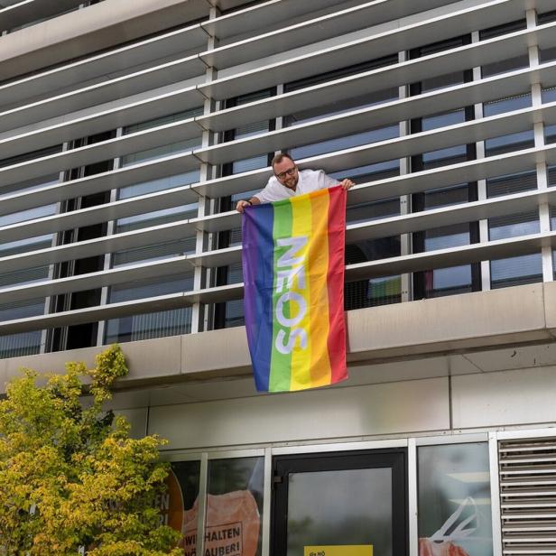 Eine Person hängt auf einem Balkon eine Regenbogenflagge mit „NEIN“ an ein modernes Gebäude.