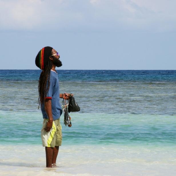 Ein Mann mit Dreadlocks und Rasta-Mütze steht im flachen türkisfarbenen Meer und hält Schmuckstücke in der Hand.