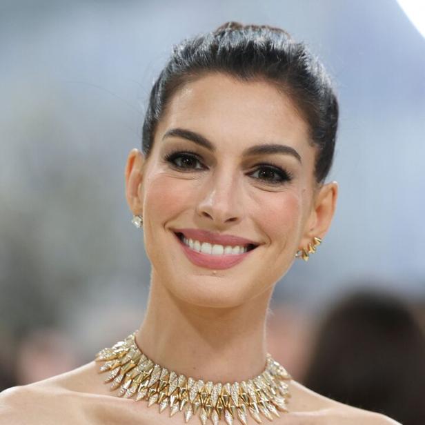 Anne Hathaway mit dunklem Haar, großem goldenen Collier und trägerlosem schwarz-weißem Kleid vor unscharfem Hintergrund.