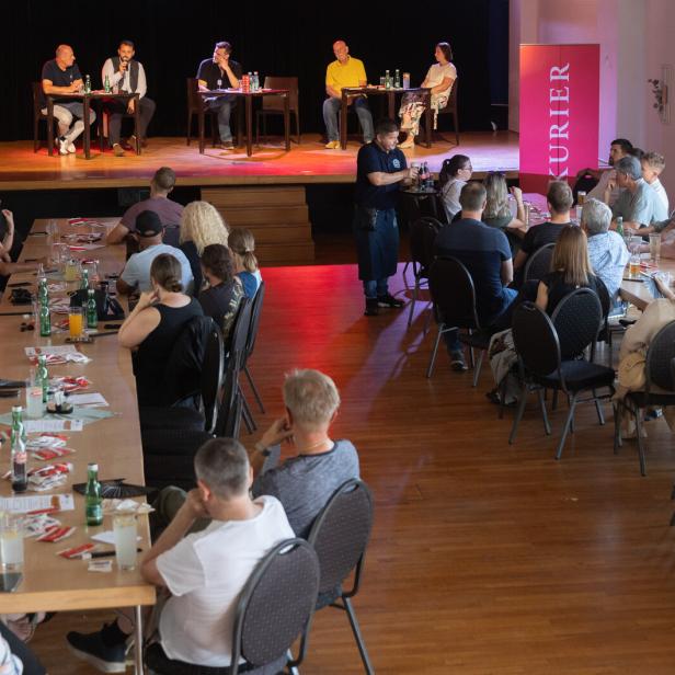 Publikum sitzt bei einer Podiumsdiskussion in einem Saal mit Bühne und roten KURIER-Bannern.