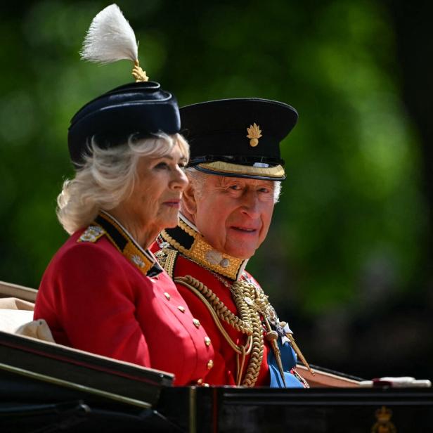 König Charles III. und Königin Camilla sitzen in einer Kutsche und tragen ihre traditionellen roten Uniformen.