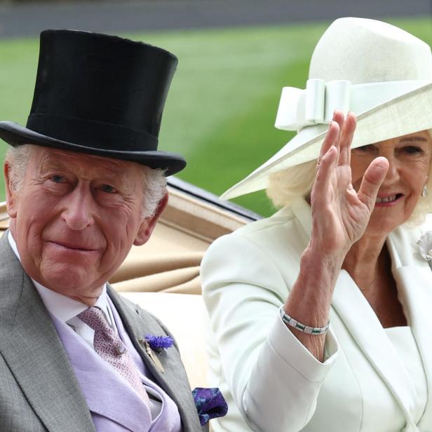 König Charles III. und Königin Camilla winken in festlicher Kleidung aus einer Kutsche.