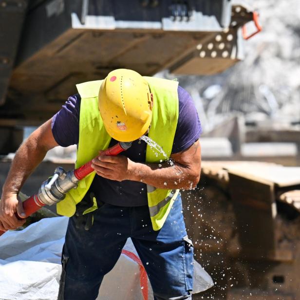 Ein Bauarbeiter mit gelbem Helm und Warnweste trinkt Wasser aus einem roten Schlauch auf einer Baustelle.