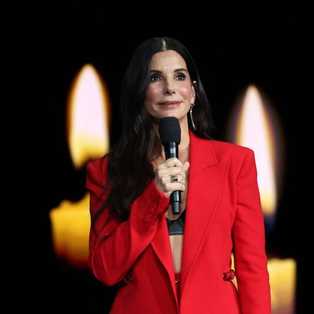Sandra Bullock spricht in einem roten Hosenanzug vor einem schwarzen Hintergrund mit unscharfen Kerzenflammen in ein Mikrofon.