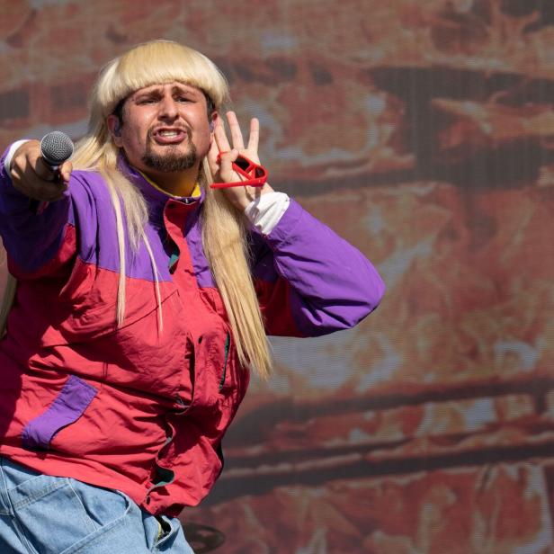 Oliver Tree mit blonder Vokuhila-Perücke und bunter Windjacke hält ein Mikrofon in die Menge und hält sich die Hand ans Ohr.
