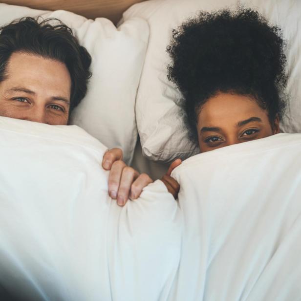 Ein lächelndes Paar liegt nebeneinander im Bett und schaut aus weißen Decken hervor.