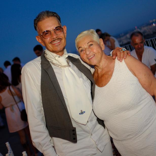 Ein Mann mit Brille und Weste sowie eine Frau in einem weißen Kleid lächeln bei einer Abendveranstaltung in die Kamera.